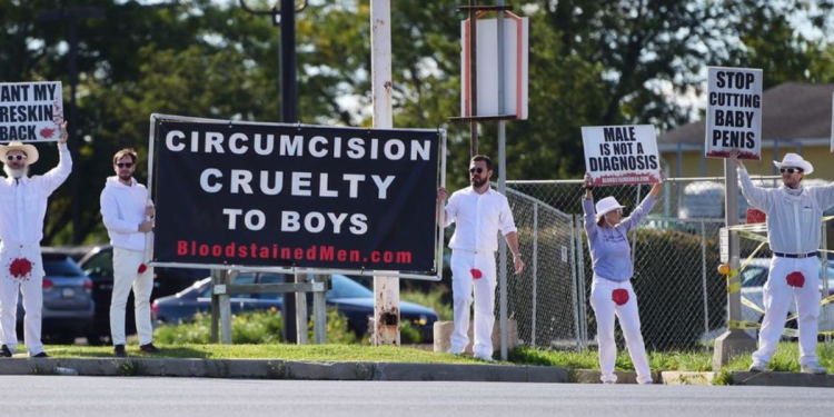 Standing outside Centura Health Primary Care in Thornton, Colorado, on April 6, two men dressed in all-white garb — except for a significant, blood-red mark on the crotch of their pants — faced traffic and held a large sign saying, "CIRCUMCISION PENIS MUTILATION."
